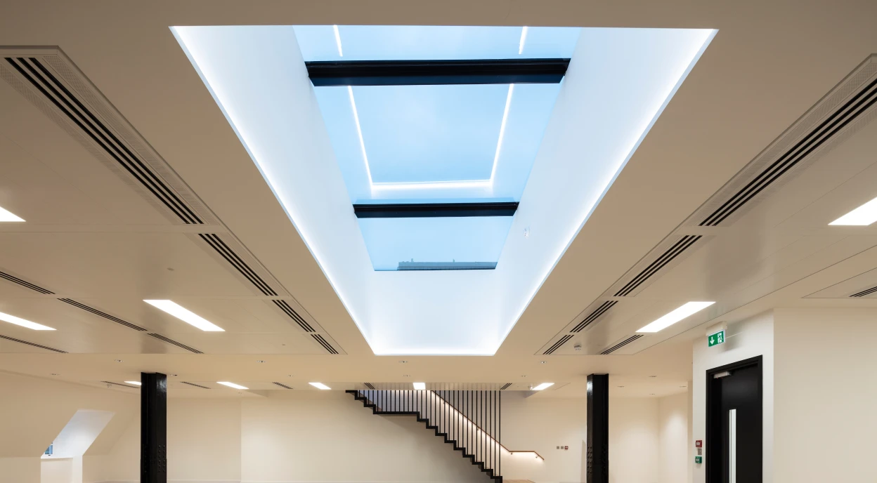Offener Raum mit hellen Wänden und einem großen Dachfenster. Dezente LED-Beleuchtung und elegante Holztreppe im Hintergrund. Der Boden ist aus Holz mit einem Fischgrätenmuster.