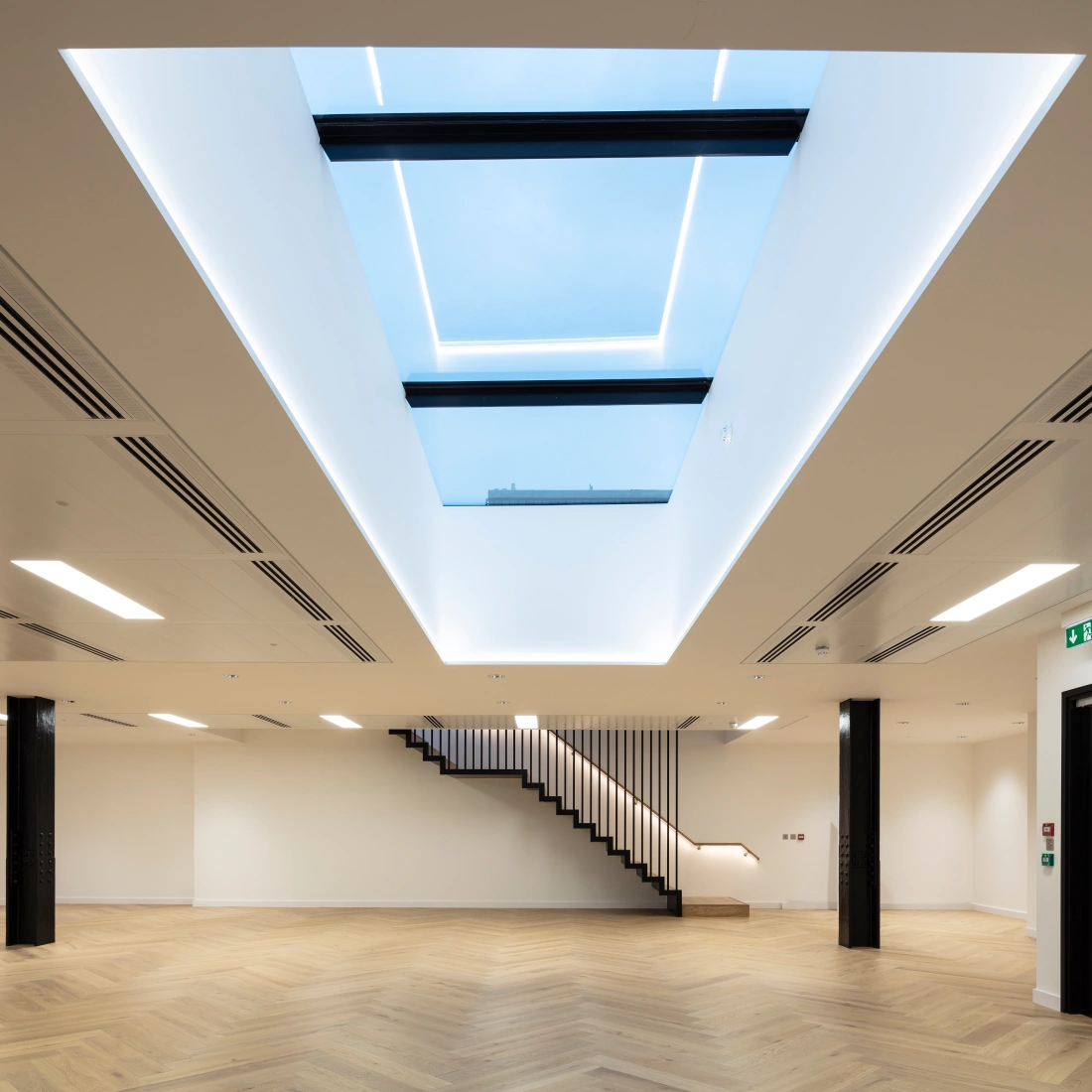 A modern, spacious room with light hardwood flooring and minimalist design. A large skylight in the ceiling allows natural light to enter, and a sleek staircase is visible in the background. Walls are painted white, enhancing the bright and airy atmosphere.