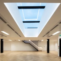 Intérieur moderne avec un plafond lumineux et un puits de lumière. Un escalier en bois blanc mène à un niveau supérieur. Le sol est en parquet clair et les murs sont peints en blanc.