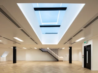 Espacio interior moderno con suelos de madera y una escalera elegante. Hay una claraboya grande en el techo que permite la entrada de luz natural. Las paredes son claras y el ambiente es minimalista.