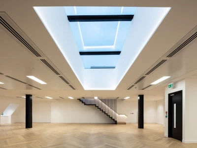 A modern, spacious room with light hardwood flooring and minimalist design. A large skylight in the ceiling allows natural light to enter, and a sleek staircase is visible in the background. Walls are painted white, enhancing the bright and airy atmosphere.