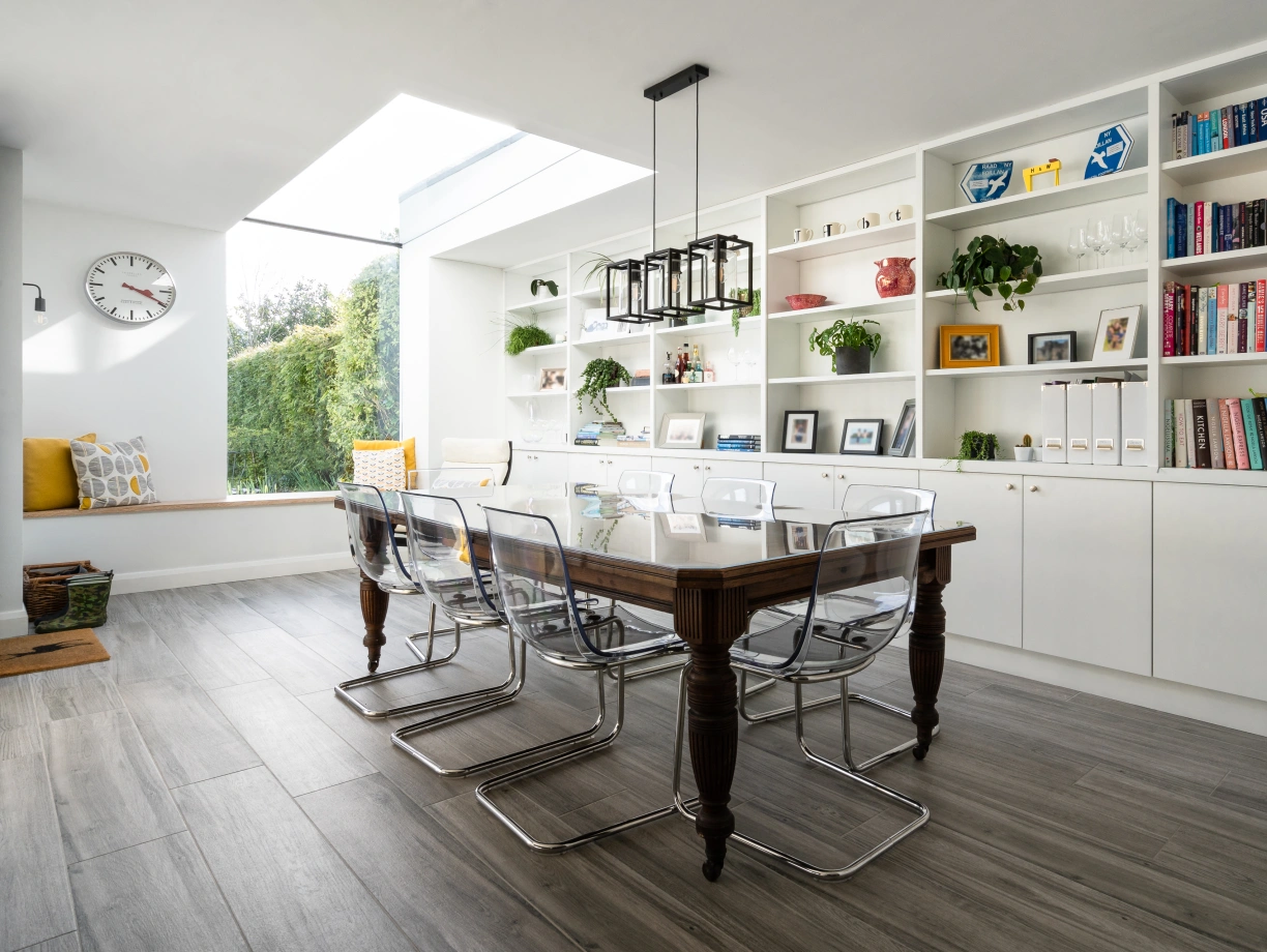 A modern dining area featuring a large wooden table surrounded by transparent chairs. There's a sleek light fixture hanging above the table. The walls have built-in shelves displaying books and decorative items, with a large window letting in natural light. A cozy seating nook with pillows is visible in the background.