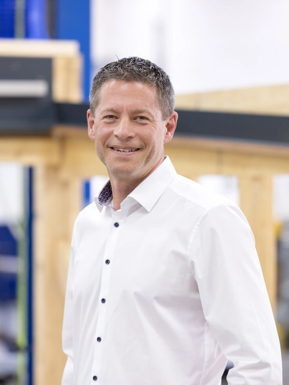 A man in a white shirt smiles confidently in a workshop setting, with wooden structures and industrial equipment visible in the background.