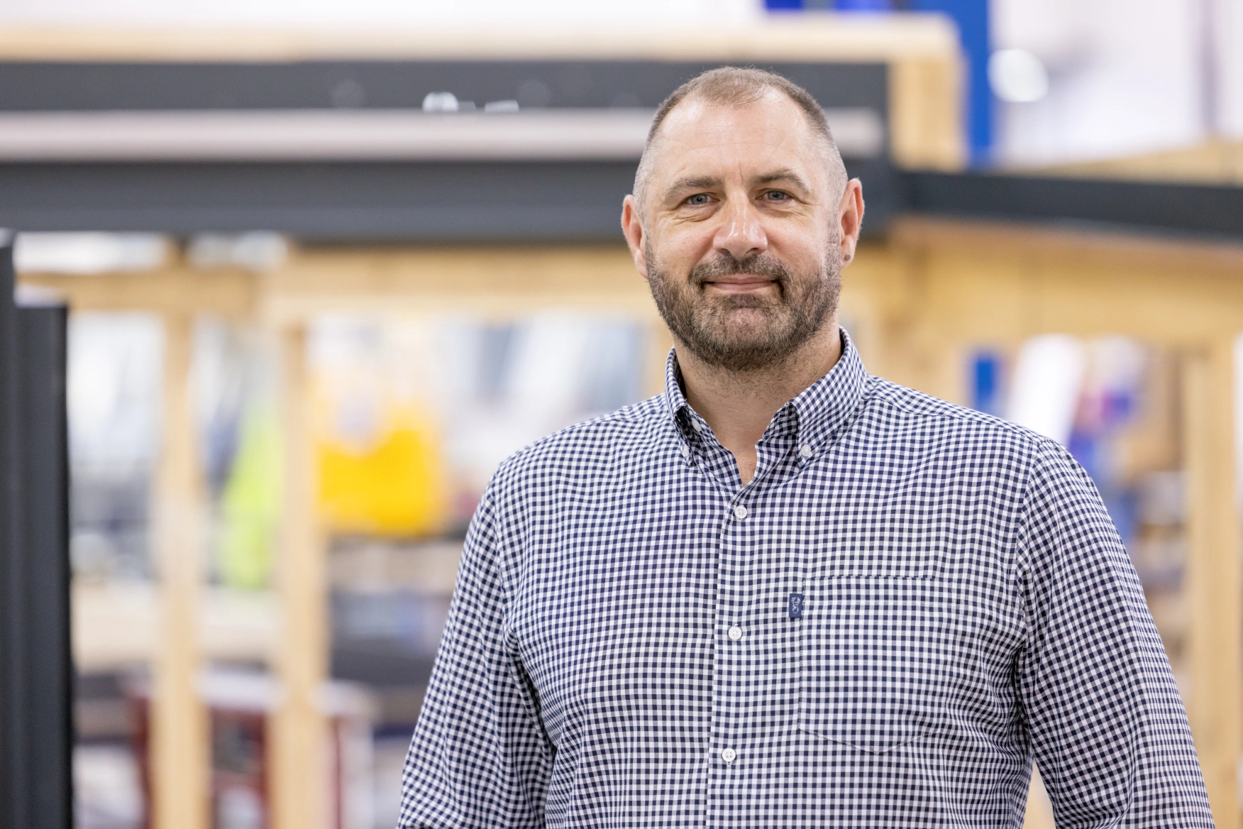 A man with a beard and short hair stands smiling in a workshop setting. He is wearing a checkered shirt and there are wooden structures and equipment in the background.