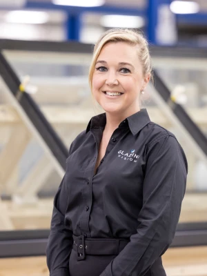 A woman in a black shirt stands smiling in front of large windows in a workshop. The background shows wooden frames and a well-lit setting.