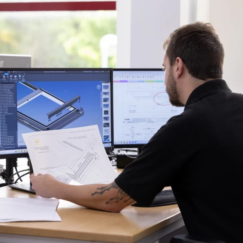 Un hombre sentado frente a un escritorio revisa un plano en papel mientras observa la pantalla de un ordenador con un software de diseño. En el fondo se pueden ver varias pantallas de computadora.