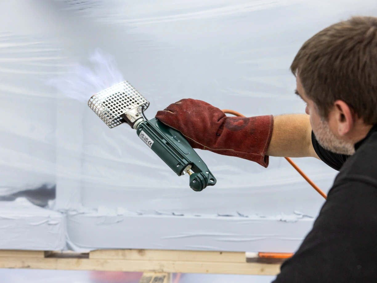 Ein Handwerker trägt Arbeitshandschuhe und verwendet ein Sprühgerät, um eine Fläche mit Farbe zu bearbeiten.rauch steigt dabei auf.