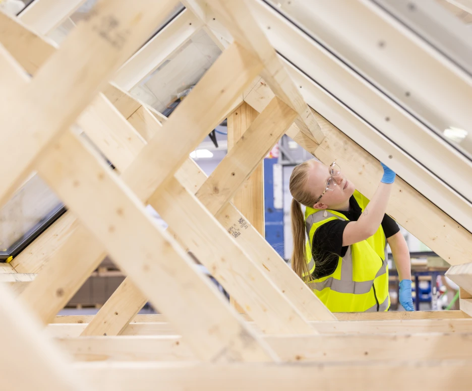 A worker in a high-visibility vest examines the frame of a structure, adjusting a component while standing inside a building construction site. The wooden beams are visible in the background, showcasing the framework of the project.