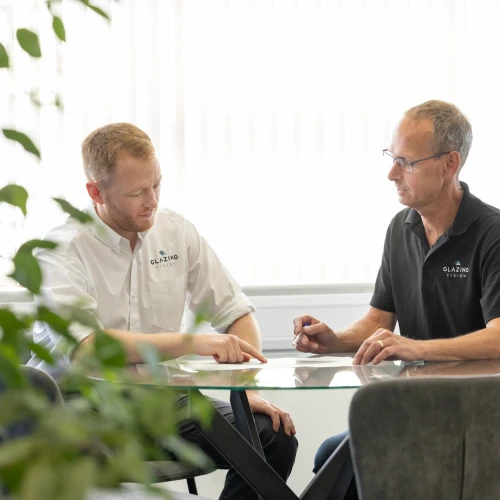 Zwei Männer in besprechender Haltung sitzen an einem Tisch im Büro. Der Mann links trägt ein helles Hemd und zeigt auf ein Dokument, während der Mann rechts in einem schwarzen Poloshirt Notizen macht. Im Hintergrund ist eine Pflanze zu sehen.