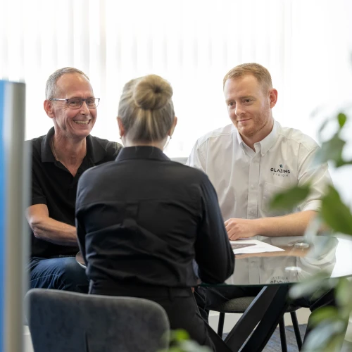 Two men and a woman are sitting around a table in a bright office. The woman, facing away from the camera, is dressed in a black shirt, while the men are engaged in conversation, smiling. One man is wearing glasses and a black shirt, and the other is in a white shirt with a logo. Green plants are visible in the foreground.