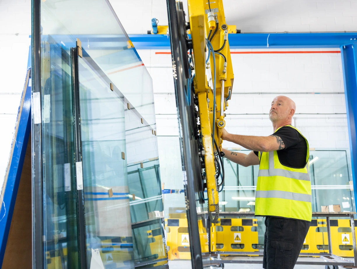 Ein Mann in einer Warnweste bedient einen Glasgreifer, um große Glasscheiben in einer Werkstatt zu handhaben. Die Umgebung ist modern mit blauen und gelben Sicherheitsbarrieren.