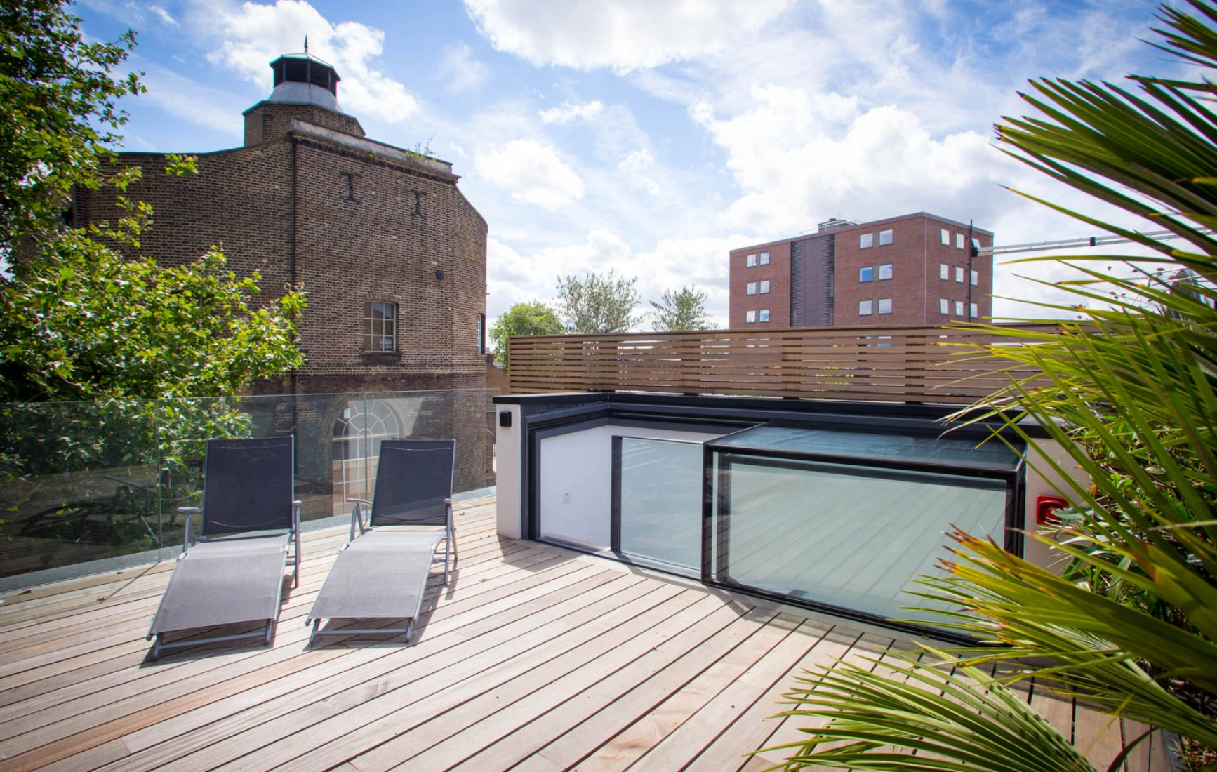 Terraza moderna de madera con dos tumbonas reclinables, rodeada de vegetación. Al fondo, se observa un edificio histórico y otro moderno bajo un cielo parcialmente nublado.