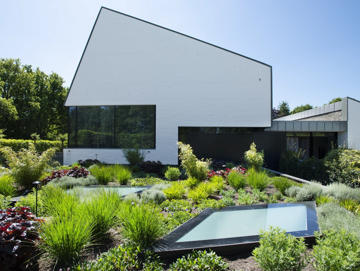 Casa moderna con una fachada blanca y un tejado en ángulo. Jardín bien cuidado con plantas verdes y áreas con agua. Cielo despejado y soleado.