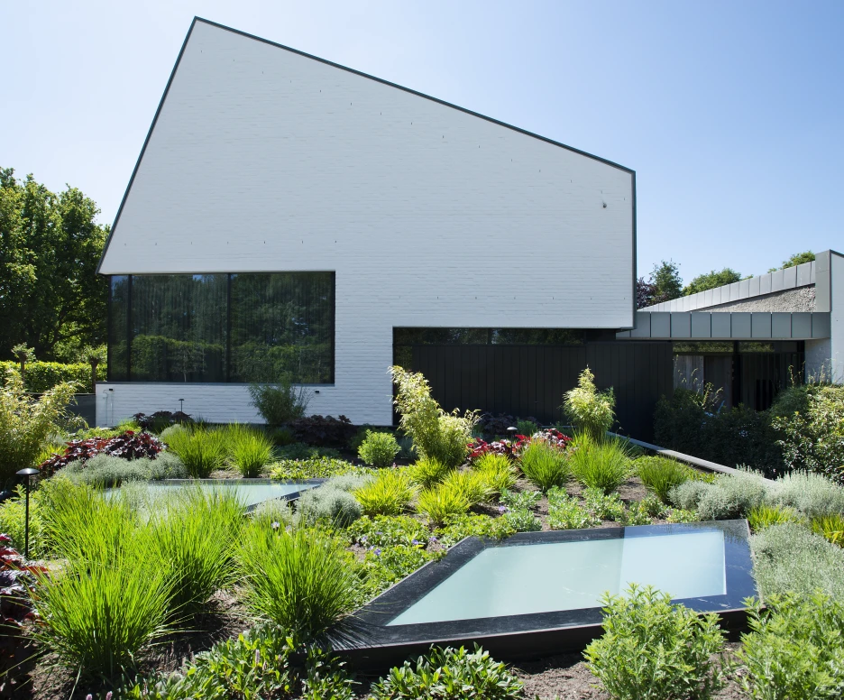 Une maison moderne à l’architecture angulaire, avec des murs blancs et de grandes fenêtres. Le jardin est verdoyant, rempli de plantes, d’herbes et de bassins d’eau rectangulaires. Le ciel est dégagé et ensoleillé.