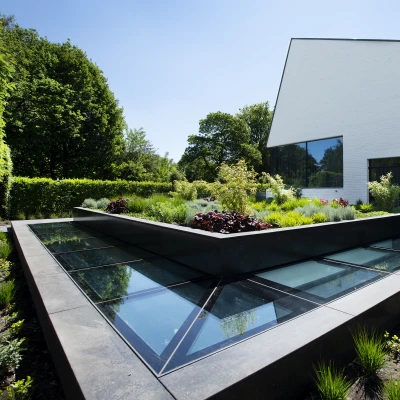 A modern building with a unique angular design is surrounded by a landscaped garden featuring lush greenery and colorful plants. The garden includes a geometric raised bed with glass panels.