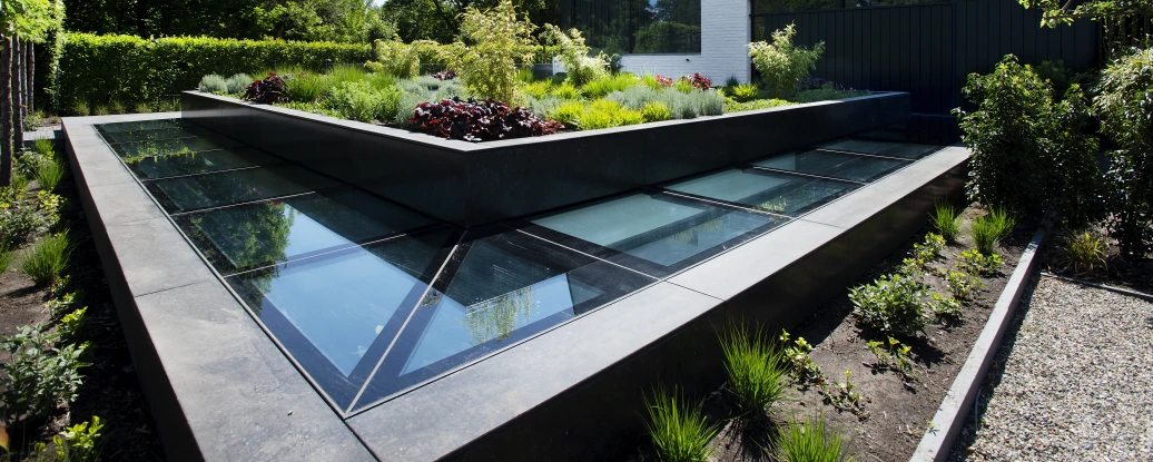 A modern building with a unique angular design is surrounded by a landscaped garden featuring lush greenery and colorful plants. The garden includes a geometric raised bed with glass panels.