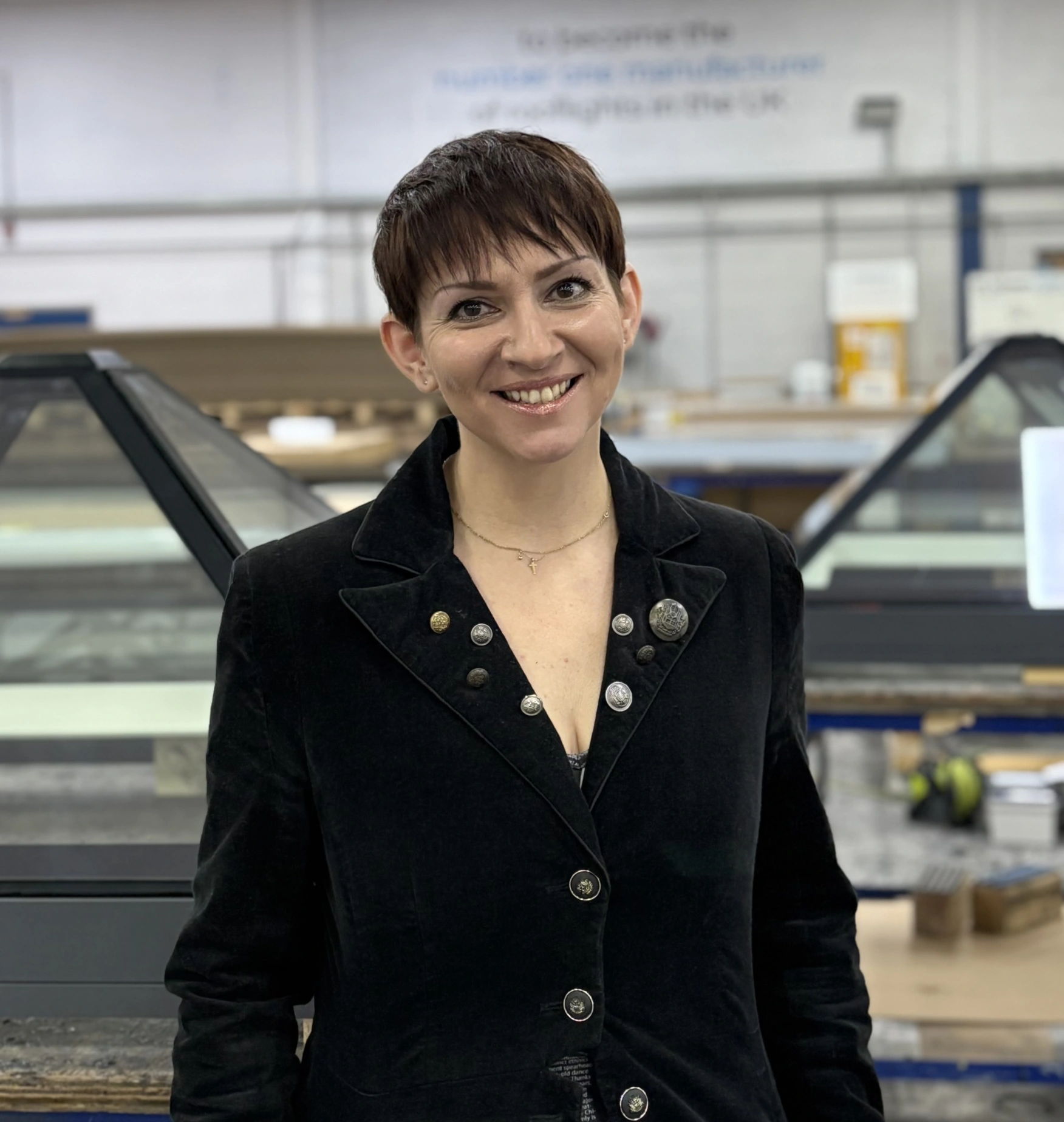 A woman with short, dark hair smiles confidently while wearing a black suit jacket. She stands in an industrial setting with glass structures and equipment in the background.