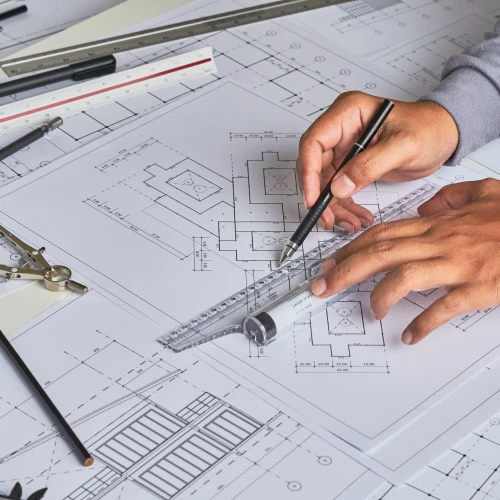 Mano sosteniendo una regla y un lápiz sobre planos arquitectónicos, rodeado de herramientas de dibujo como compás y lápices.