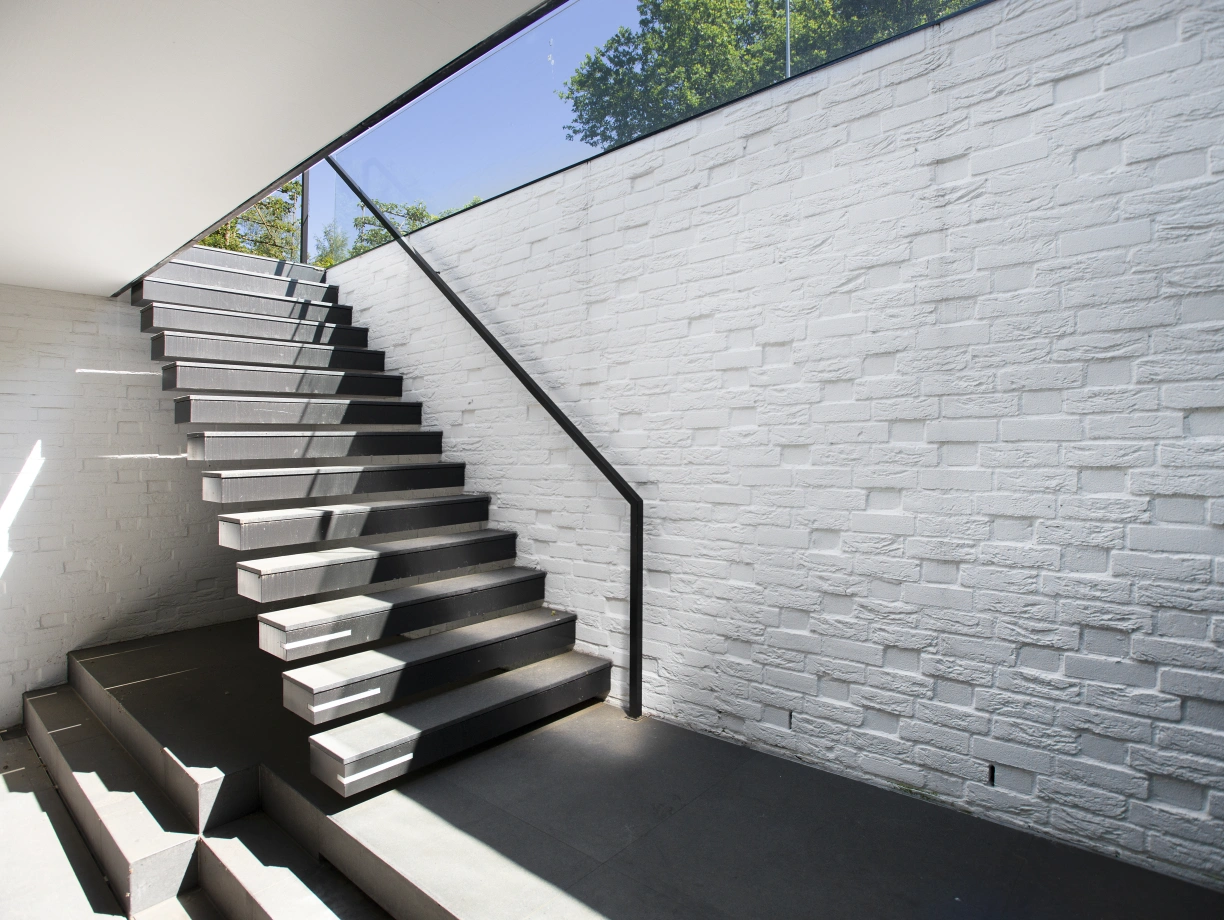 Un escalier moderne avec des marches noires et un garde-corps en verre, menant vers un espace lumineux, contre un mur en briques blanches. La lumière naturelle éclaire l'escalier.
