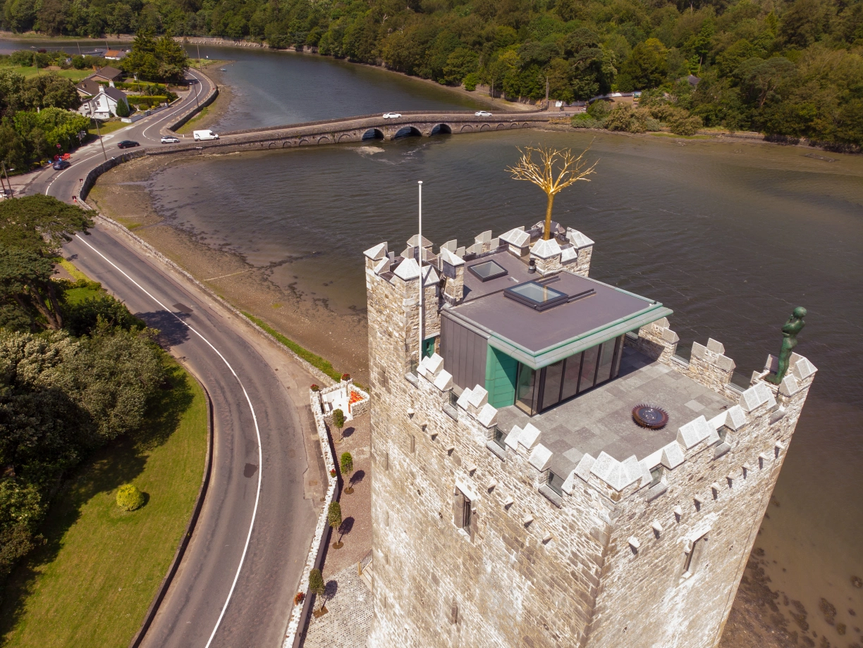 Aerial view of a stone tower with a modern glass structure on top, located near a river. Surrounding greenery and a road alongside the water are visible. A bridge crosses the river in the background.