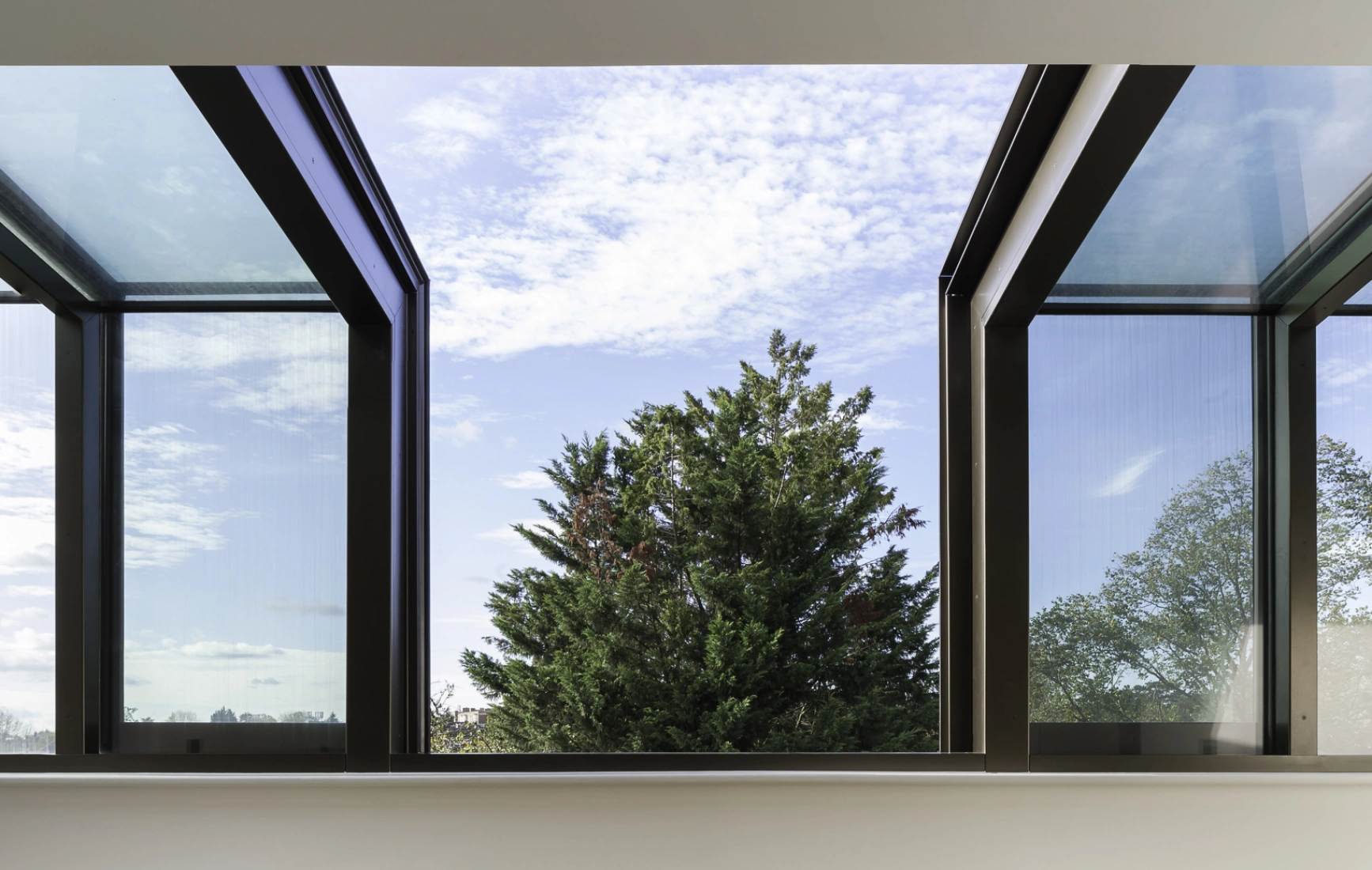 Large windows with a modern design open up to a view of a green tree and blue sky with scattered clouds. The interior features light-colored walls, wooden flooring, and minimalist furniture.