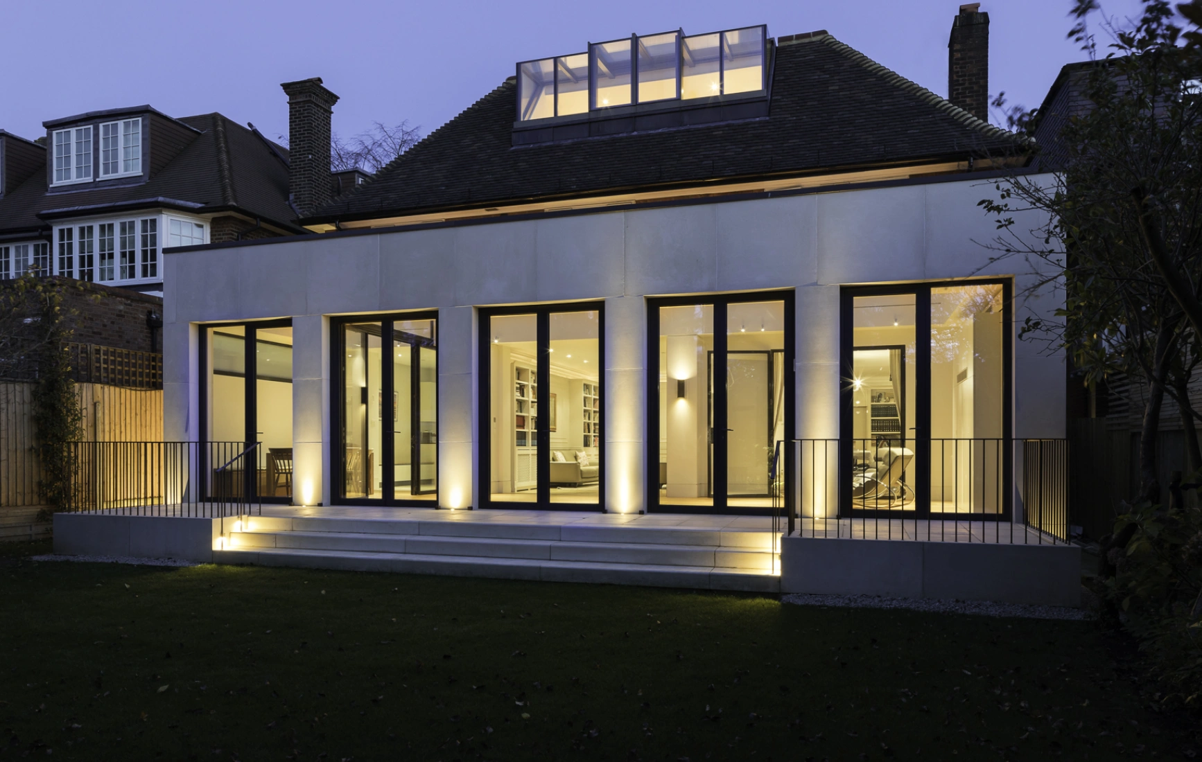 Maison moderne avec de grandes fenêtres illuminées au crépuscule. Le jardin est soigné, entouré de clôtures en bois. Un toit en pente et un vitrage au-dessus de la porte ajoutent du caractère.