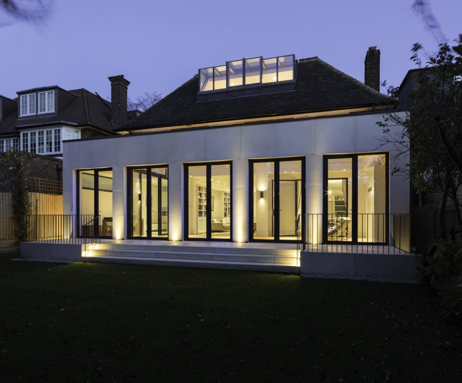 Maison moderne avec de grandes fenêtres illuminées au crépuscule. Le jardin est soigné, entouré de clôtures en bois. Un toit en pente et un vitrage au-dessus de la porte ajoutent du caractère.