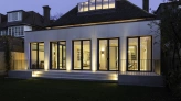 A modern house at dusk, featuring large glass windows that illuminate the interior. The structure is surrounded by a green lawn and has a wooden fence in the background. The roof has a skylight, adding to its contemporary design.