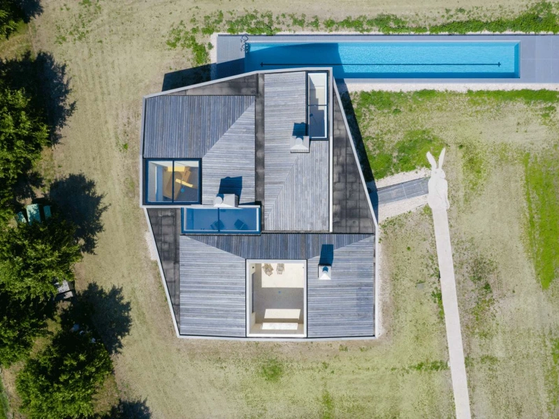 Casa moderna con diseño angular y terraza, rodeada de césped. Una piscina alargada se encuentra al lado del edificio.