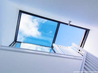 Blick nach oben durch ein großes Glasdach, das blauen Himmel und Wolken zeigt, mit einer Treppe im Vordergrund.