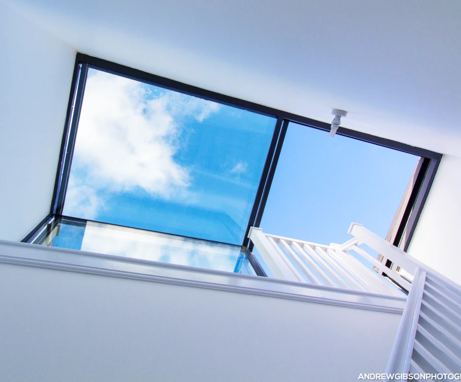 Vue vers le ciel à travers un plafond en verre, avec des nuages blancs et un éclairage naturel. Un escalier blanc mène vers le haut.
