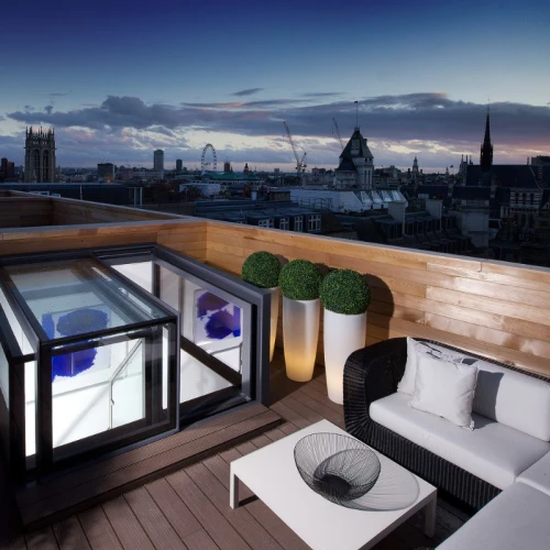 Terraza moderna con sofá blanco y mesa de café, decorada con plantas en macetas, vista panorámica de la ciudad al atardecer con nubes y edificios.