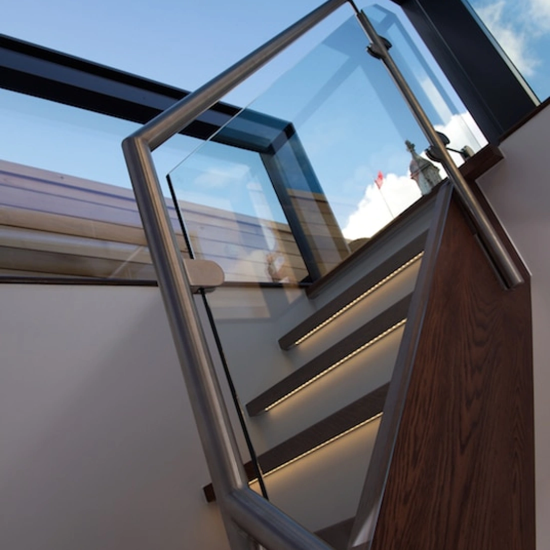A modern staircase with wooden steps and a sleek glass railing. Natural light streams in from a skylight above, illuminating the space.