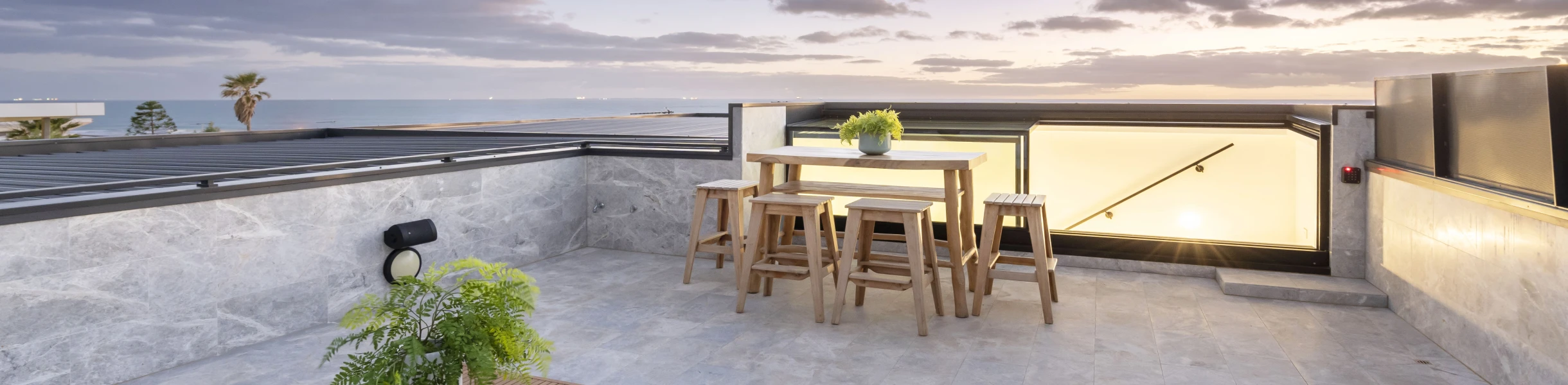 Terraza moderna con mesas y sillas de madera, decorada con plantas. Se observa un atardecer con nubes y palmeras en el horizonte.