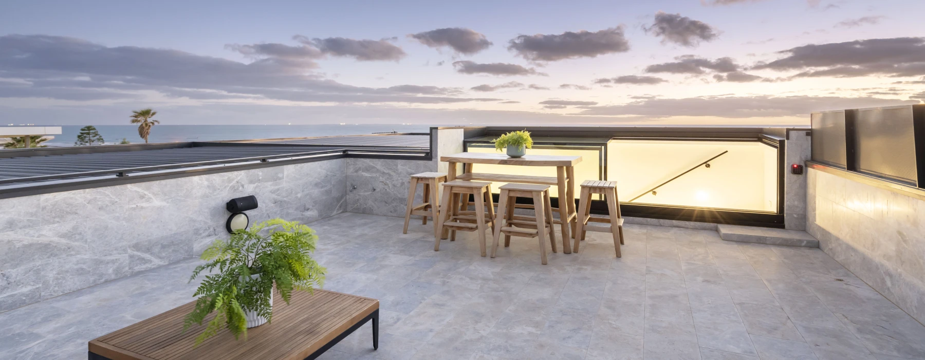 Terrasse spacieuse avec une table et des chaises en bois, décorée d'une plante en pot. Vue sur un ciel coloré au coucher de soleil avec des nuages. Un palmier est visible au loin.