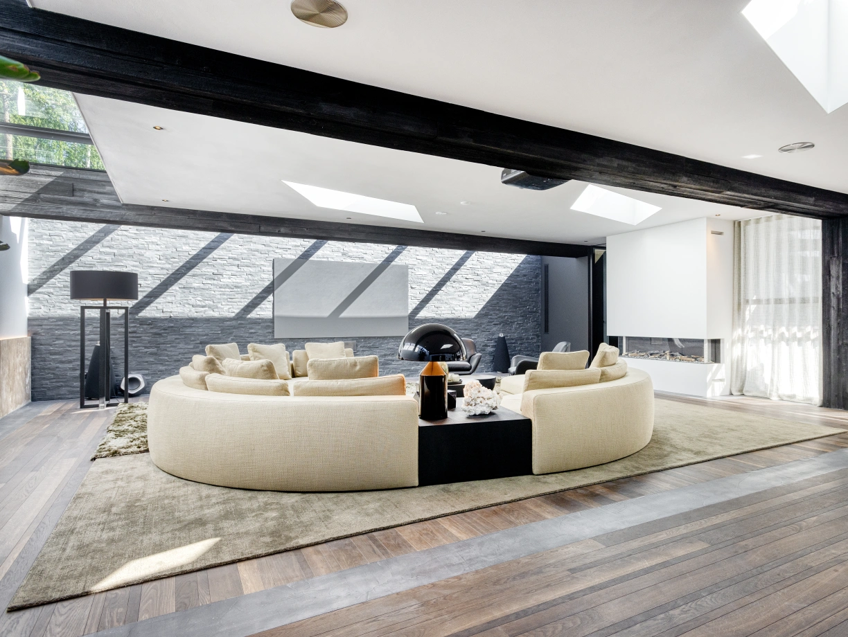 A modern living room features a large round beige sofa arranged in a circle, surrounded by hardwood flooring. Skylights illuminate the space, highlighting a unique black and white decor. A small coffee table sits in the center, with a stylish lamp and decorative items.
