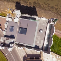 Aerial view of a stone castle with a modern flat roof, situated near a body of water. The landscape includes a winding road, green grass, and a tree with golden branches.