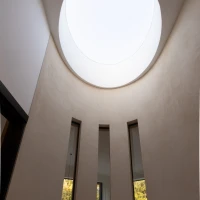 Runder Innenraum mit einer großen, kreisförmigen Öffnung an der Decke, die Licht hereinlässt. Drei vertikale Fenster an den Wänden bieten Ausblicke ins Freie. Die Wände sind in einem hellen, neutralen Farbton gehalten.