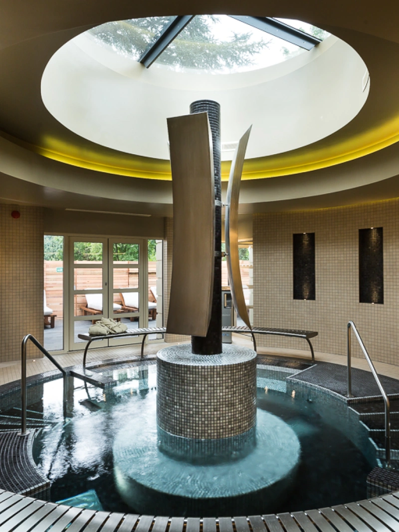 Un espace de spa moderne avec une grande piscine circulaire en mosaïque, entourée de murs en tuiles claires. Des colonnes métalliques élégantes s’élèvent au centre, tandis qu’un toit en verre laisse passer la lumière. Des chaises longues sont visibles à l’extérieur.