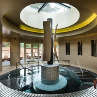 A modern spa interior features a circular skylight and a central water feature with sleek sculptures, surrounded by tiled walls. Comfortable lounge chairs are visible through large glass doors leading outside. Plush towels are neatly arranged on the side.