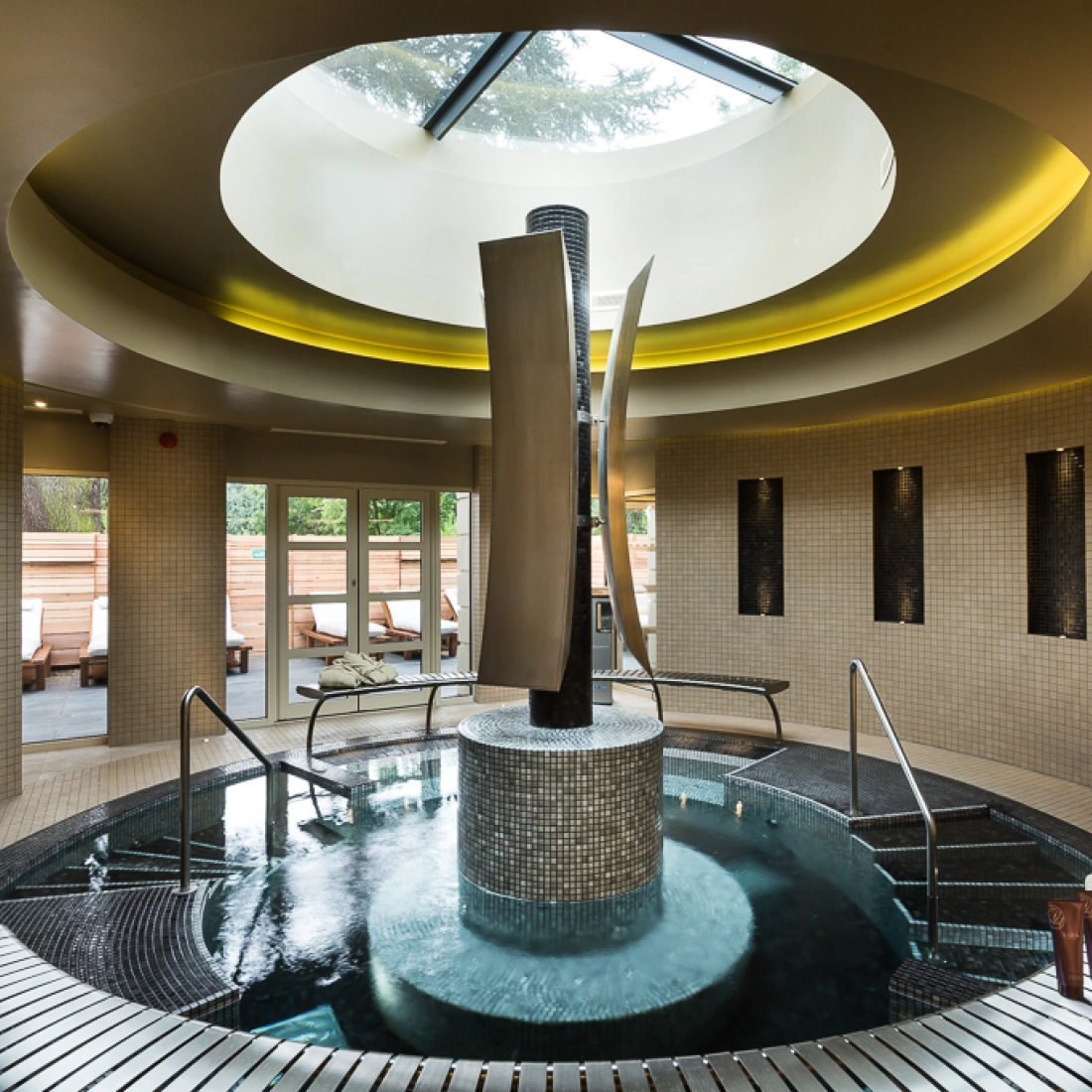 Spa moderno con una fuente central y jacuzzi. La iluminación suave resalta las paredes de azulejos claros y las ventanas permiten la entrada de luz natural. Toallas enrolladas y un ambiente relajante completan la escena.