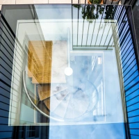 Ein runder Treppenaufgang aus Metall, sichtbar durch ein großes, transparentes Glasdach. Über dem Treppenhaus spiegeln sich Wolken im Glas, während die Seiten mit schwarzen Gitterelementen umrahmt sind. Ein Holztisch steht im oberen Bereich.
