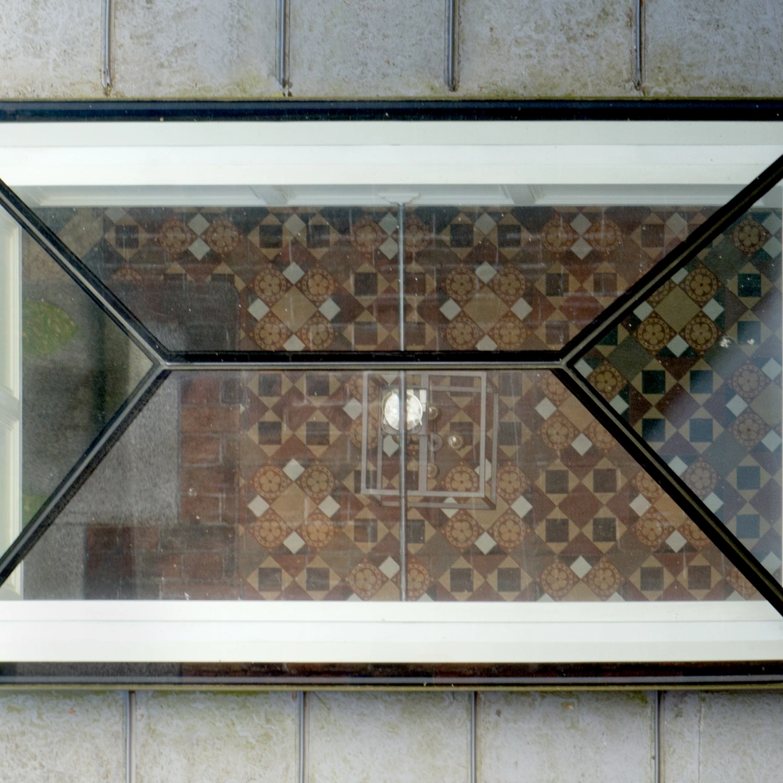 A geometric window framed in black, reflecting a patterned tiled floor with intricate designs in various colors. Natural light illuminates the space visible through the window.