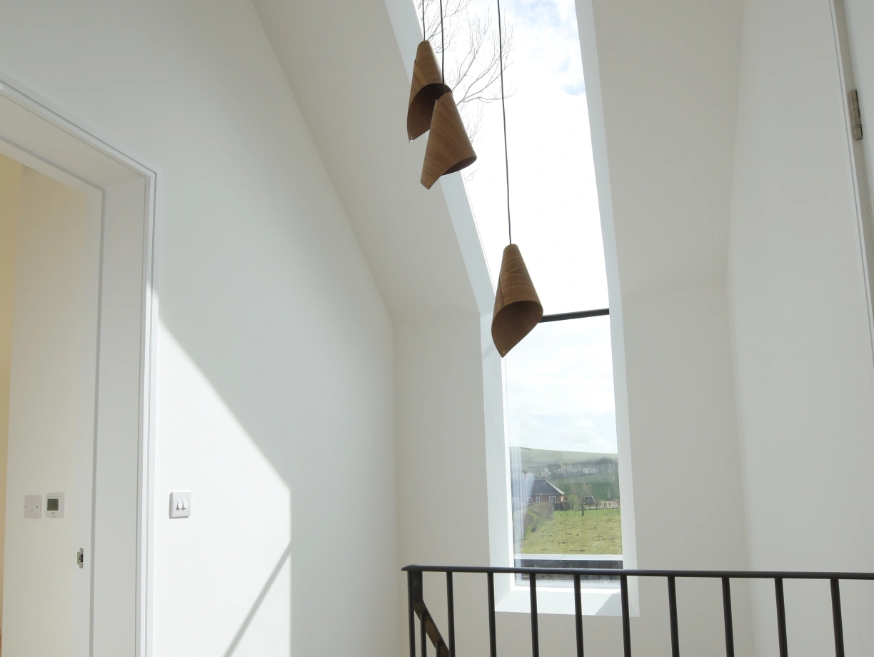 A bright hallway with a modern design, featuring a large skylight and three pendant lights hanging from the ceiling. There is a black railing along the staircase leading down, and the walls are painted white, creating a clean and airy atmosphere.