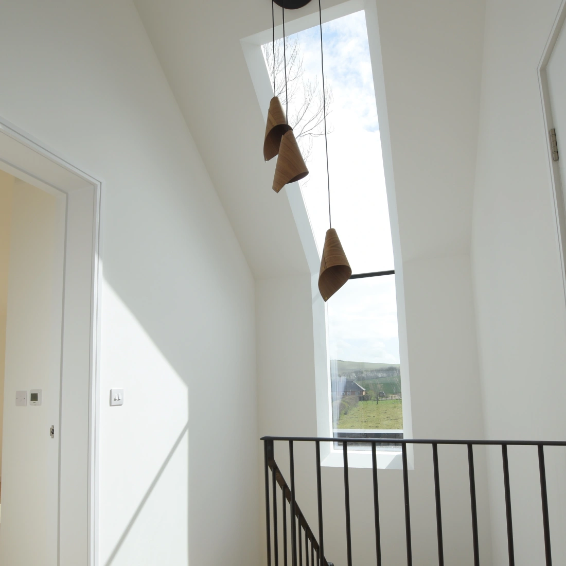 Helle Treppe mit Holzfußboden und schwarzem Geländer. Drei hängende Lampen aus Keramik. Großes Fenster lässt Tageslicht herein. Wände und Decken sind weiß gestrichen.