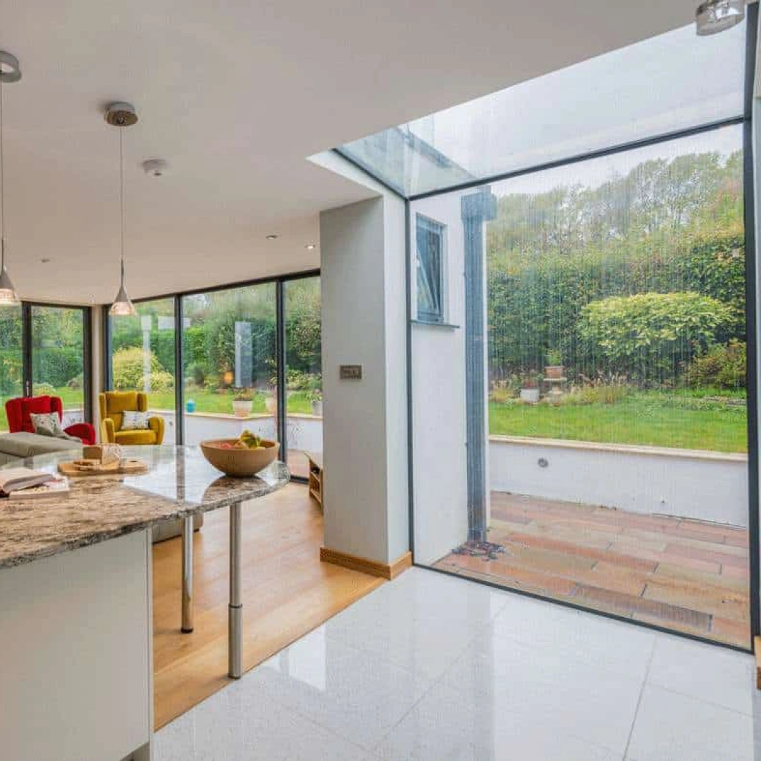 Moderne Wohnküche mit offenem Grundriss. Ein Tisch mit einer Vase von frischen Blumen steht im Vordergrund. Große Fenster bieten Aussicht auf einen Garten mit grünen Pflanzen und Rasen. Gemütliche Sitzgelegenheiten sind im Hintergrund sichtbar.