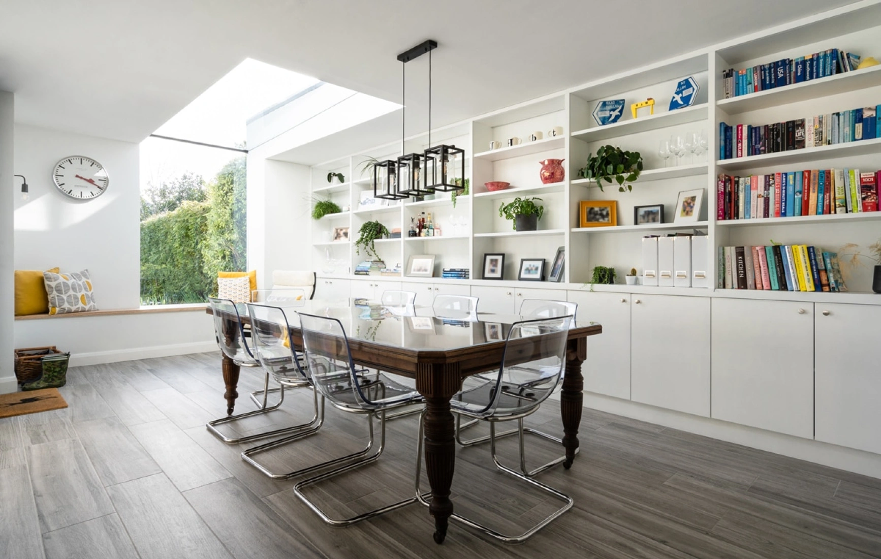 Comedor elegante con mesa de madera y sillas transparentes. Estanterías blancas llenas de libros y decoraciones. Ventana grande que ilumina el espacio. Detalles como plantas y un rinconcito con almohadas.