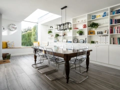 In a dining area with a table in the centre with glass chairs all around it. To the right there is a long whiite book shelf going across the whole wall which is filled with books and plants. To the left there is a sitting area with yellow cushions and a big clock above which is next to an eaves skylight.