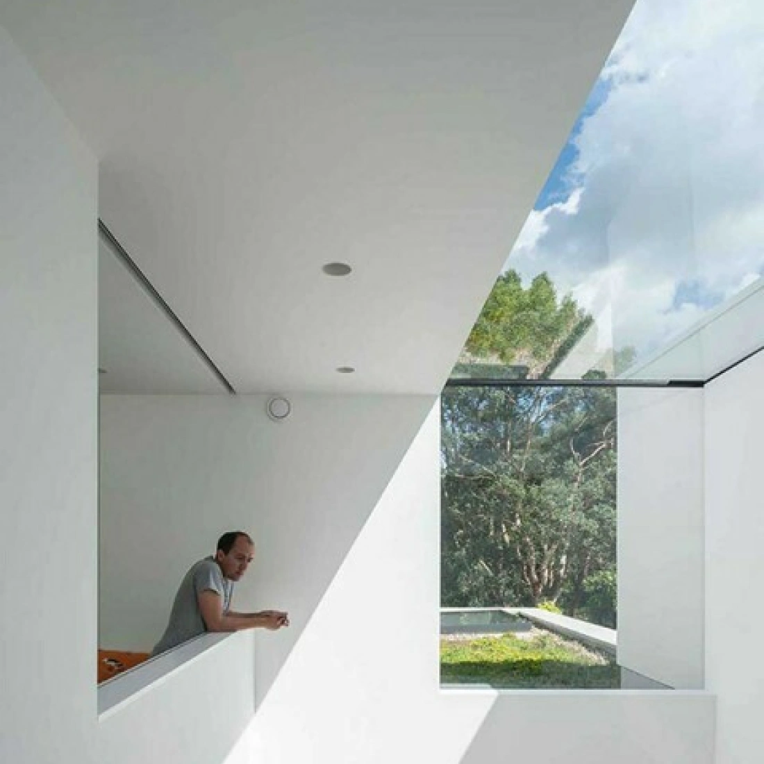 A man leans on a white railing in a modern, minimalist interior. Large windows allow natural light to flood in, showcasing a view of trees and blue sky. The geometric lines of the architecture create a sharp contrast with the organic landscape outside.