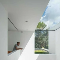 Modernes, minimalistisches Innendesign mit einem Mann, der an einem weißen Geländer lehnt. Große Fenster lassen Tageslicht herein und bieten einen Blick auf die grüne Umgebung. Geometrische Linien erzeugen eine interessante Komposition.